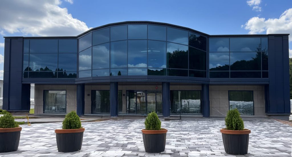 Zdjęcie przedstawia front dwupiętrowego budynku o nowoczesnej architekturze z dominującymi dużymi, ciemnymi przeszkleniami, w których odbijają się chmury. Przed wejściem znajduje się plac wyłożony jasną kostką brukową, na którym ustawiono cztery ciemne donice z przystrzyżonymi, zielonymi krzewami.